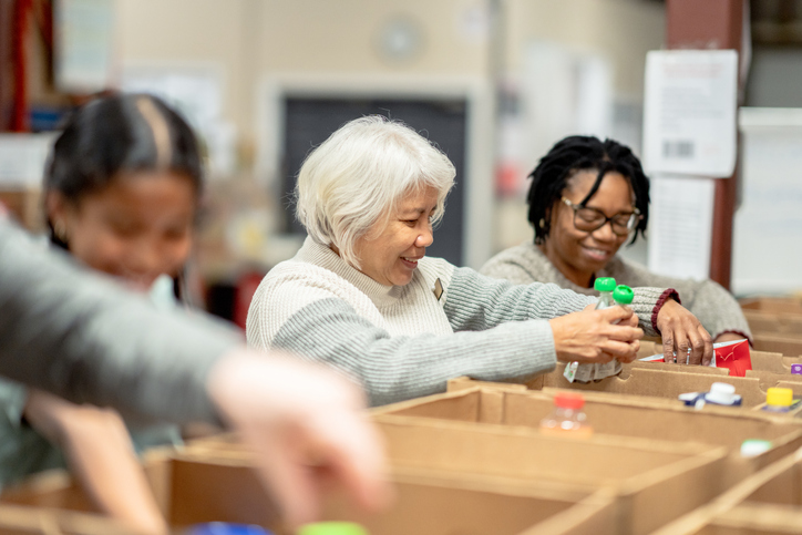 A small group of four volunteers are seen standing at a table full of cardboard boxes as they pack them with non-perishable food items at a local Food Bank. A young mixed race girl is working with three seniors as they happily give of their time for the cause and carefully pack the items into the boxes.